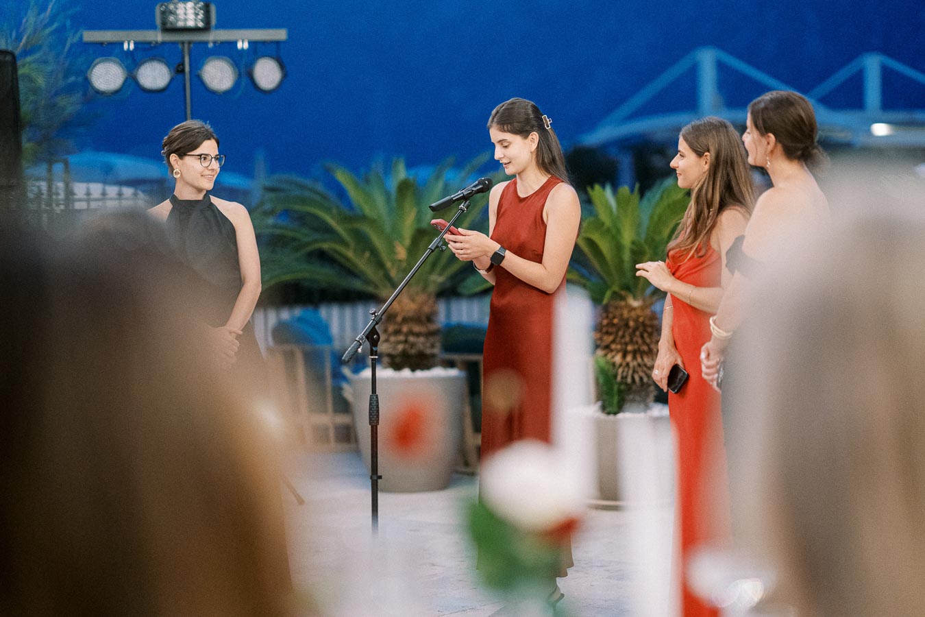 Four women in elegant dresses engage in a speech at an outdoor event with tropical plants in the background.