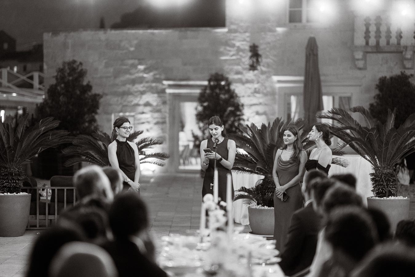 Black and white image of an elegant outdoor evening event with a group of formally dressed women standing by a microphone, delivering a speech to seated guests.