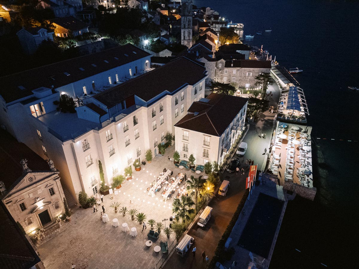Aerial view of a picturesque coastal town at night, featuring illuminated buildings, a vibrant waterfront promenade, and a gathering in a well-lit courtyard, creating a lively and inviting atmosphere.