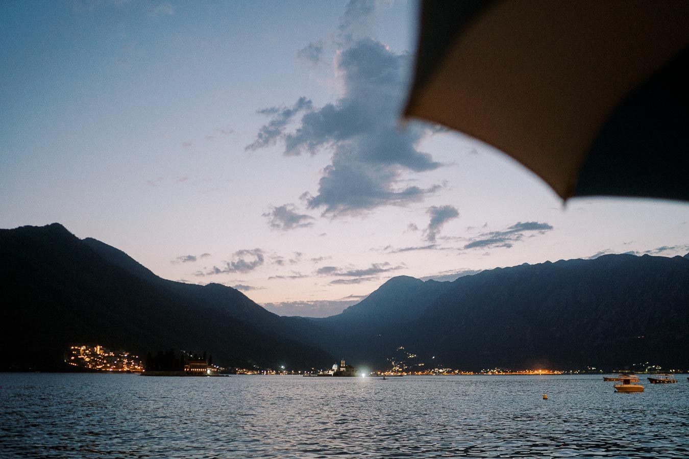 Scenic view of a serene coastal village at dusk with mountains in the background, illuminated by soft evening lights and framed by an umbrella in the foreground.