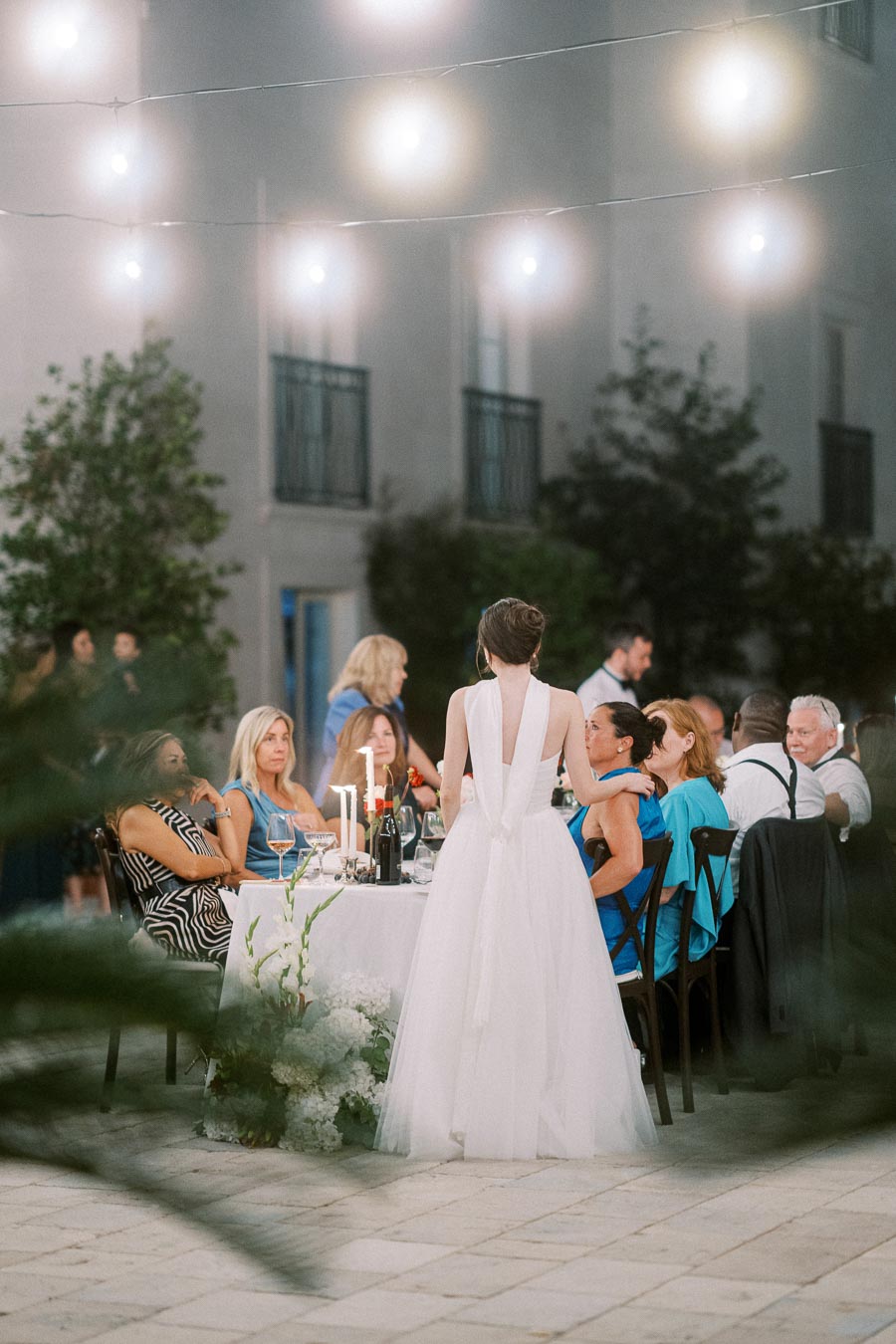 Outdoor wedding reception with guests seated at a candlelit table, soft lighting above, and a woman in a white dress interacting with attendees in an elegant evening setting.
