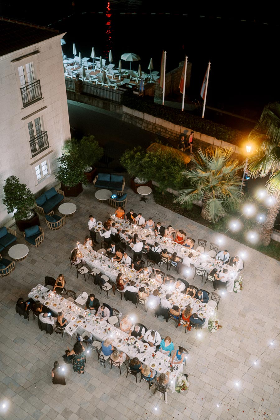 Aerial view of an elegant outdoor dinner party at night, with guests seated at long tables adorned with white tablecloths and decorative lights, set on a stone courtyard near a waterfront area with beach chairs and umbrellas.