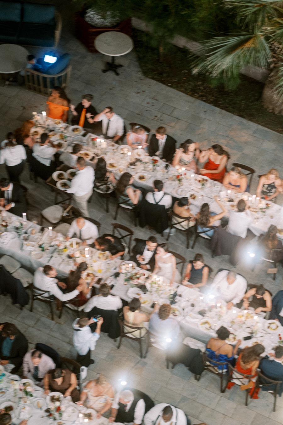Aerial view of a large, elegant wedding reception dinner with guests seated around long, candlelit tables set outdoors on a stone patio, surrounded by greenery.