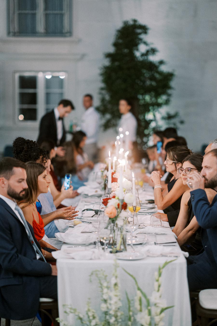 Elegant evening wedding reception with guests seated at a long, candlelit dinner table adorned with floral arrangements.