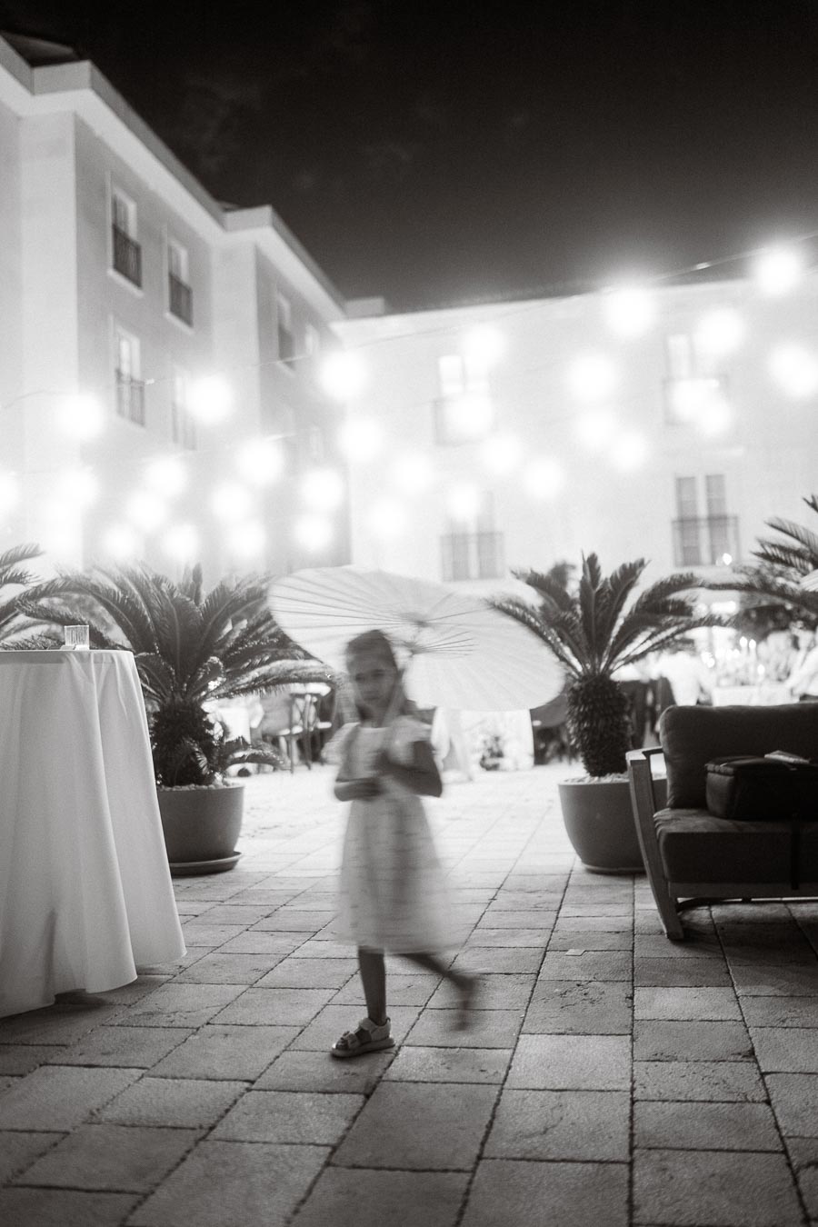 A blurred black and white photo of a young girl holding a parasol, walking on a cobblestone patio at night, surrounded by soft, glowing lights and potted plants in an elegant outdoor setting.