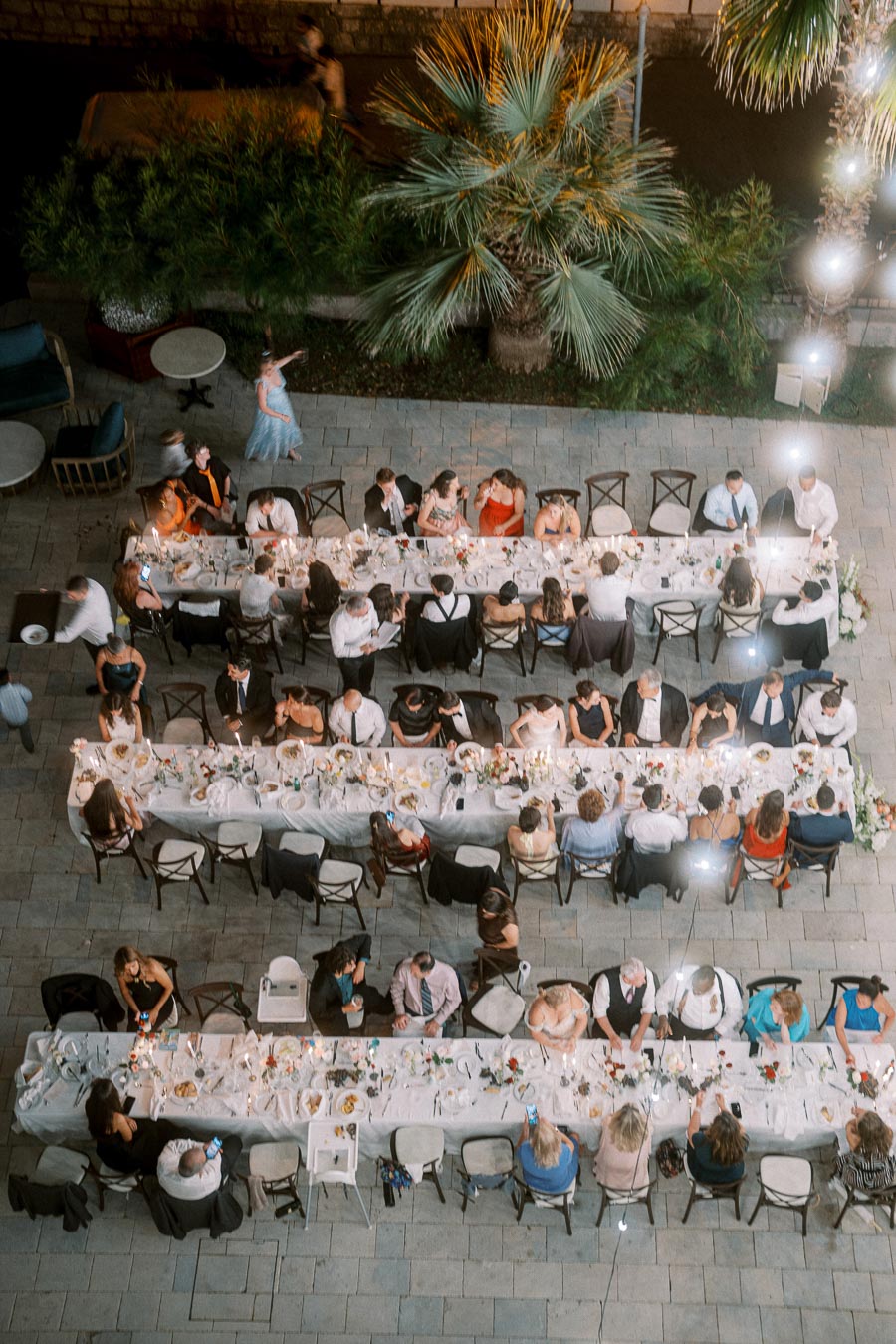 Aerial view of an elegant outdoor wedding reception with long banquet tables, surrounded by guests in formal attire, under soft evening lighting, with lush greenery in the background.