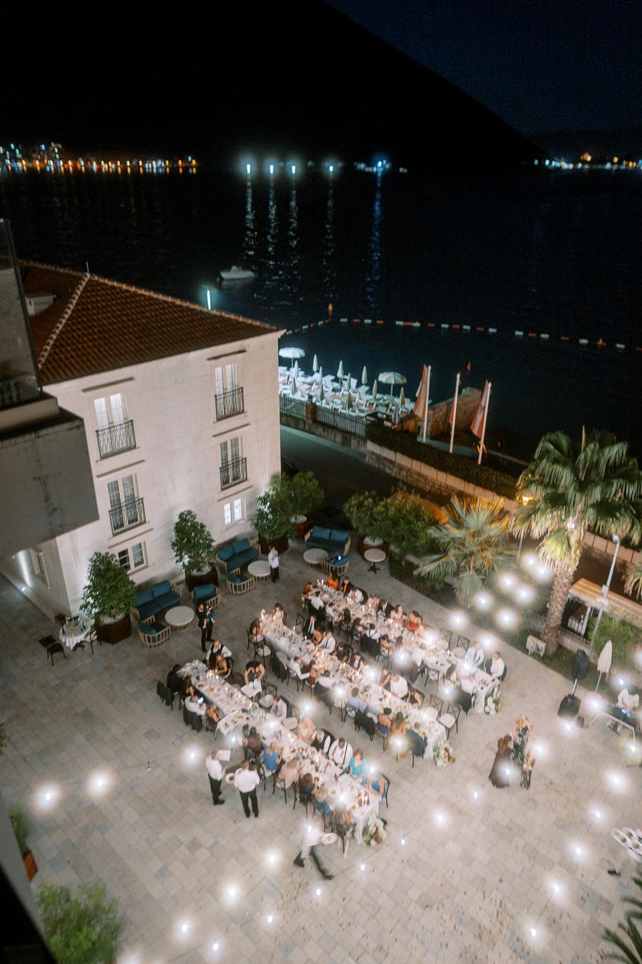 Aerial view of an elegant outdoor dinner party by the waterfront at night, featuring long tables with guests enjoying a meal next to a scenic building and palm trees, with reflections of city lights shimmering on the water in the background.