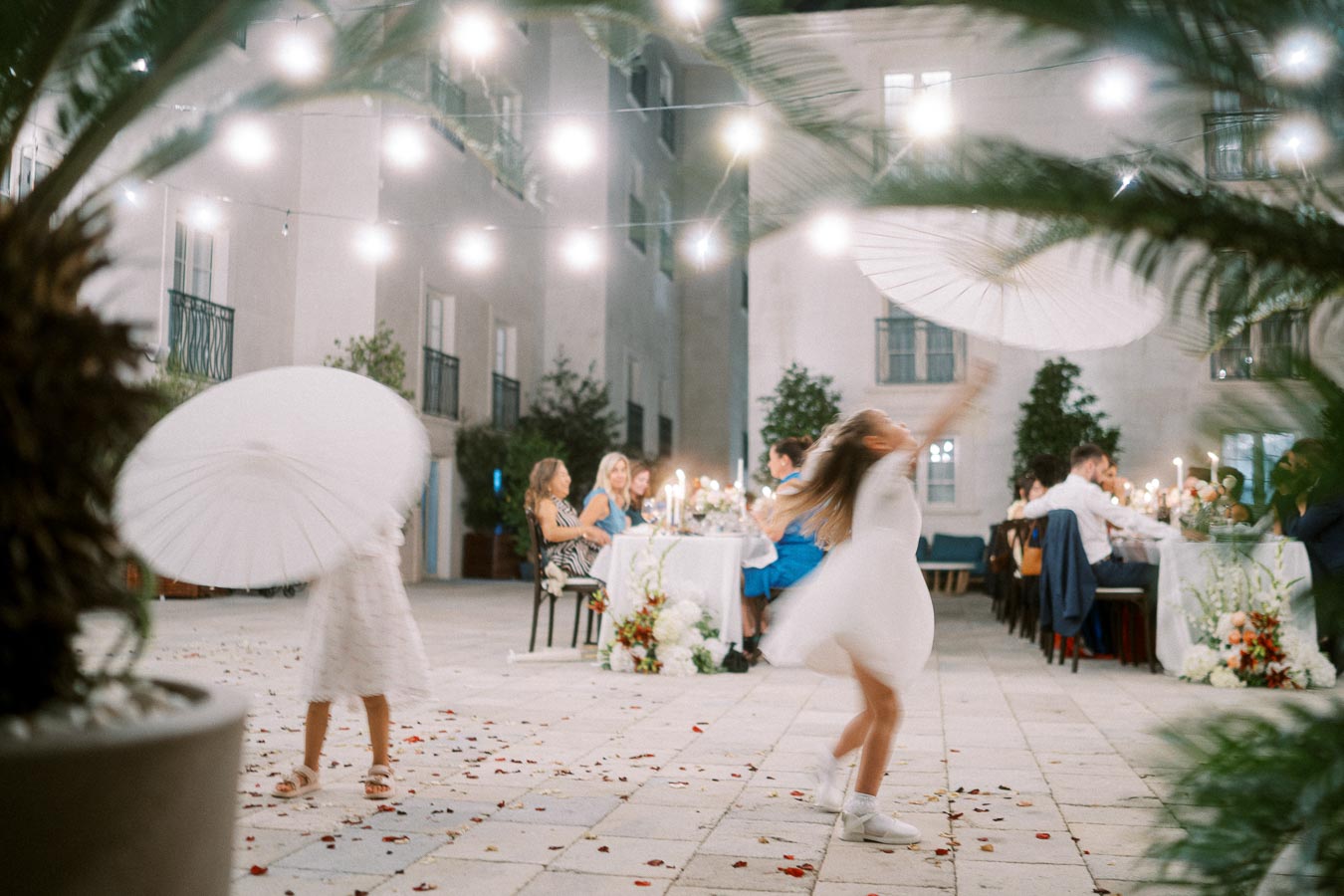 Outdoor evening wedding reception with children playfully dancing with white umbrellas, string lights illuminating guests at a beautifully decorated dinner table.