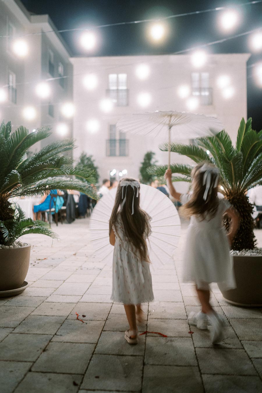 Two young girls in white dresses holding parasols, walking in a beautifully lit courtyard with string lights and lush potted plants, creating a whimsical and festive evening atmosphere.