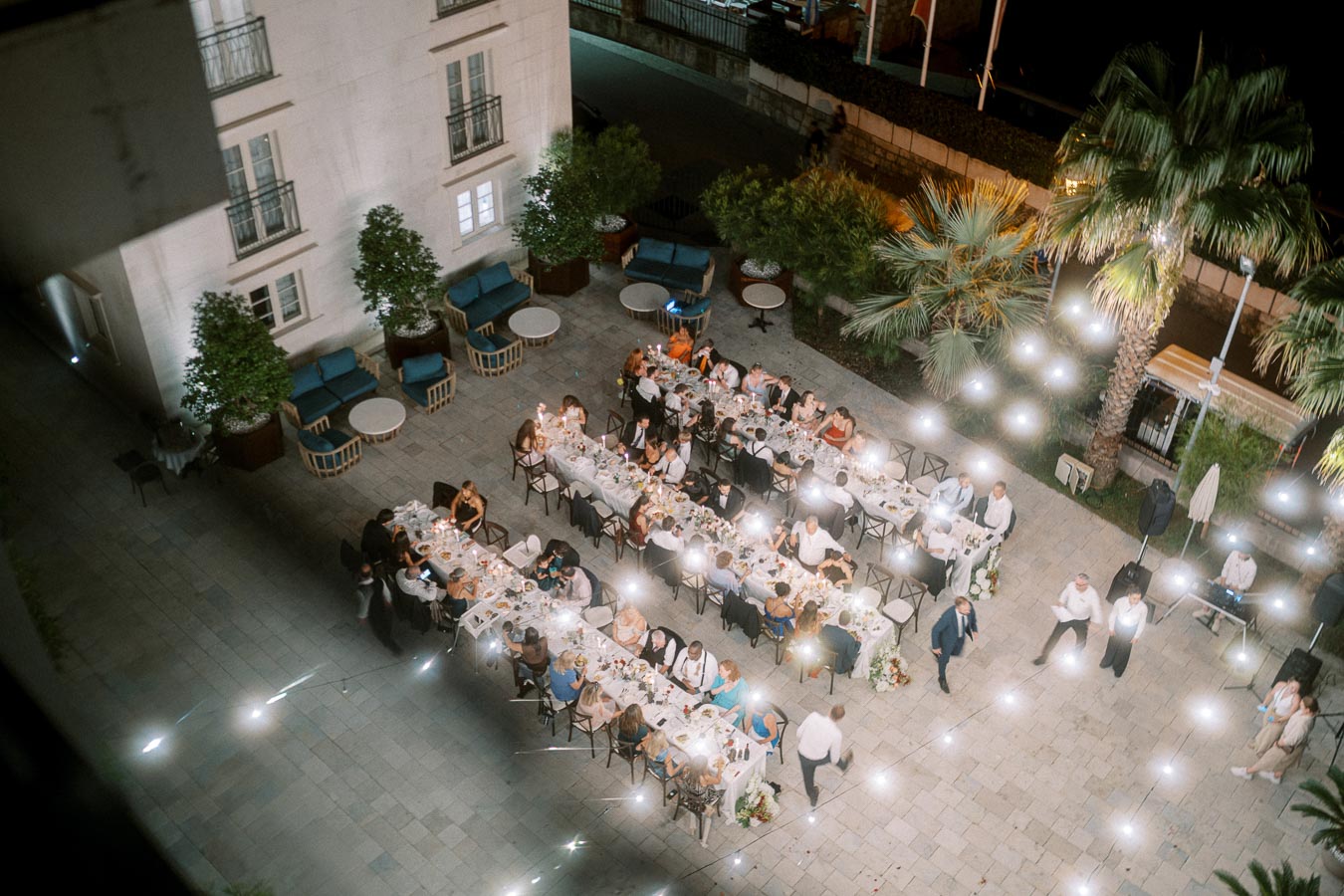 Aerial view of an elegant outdoor evening party with a long dining table surrounded by guests, decorated with string lights, located in a courtyard with palm trees and modern seating.