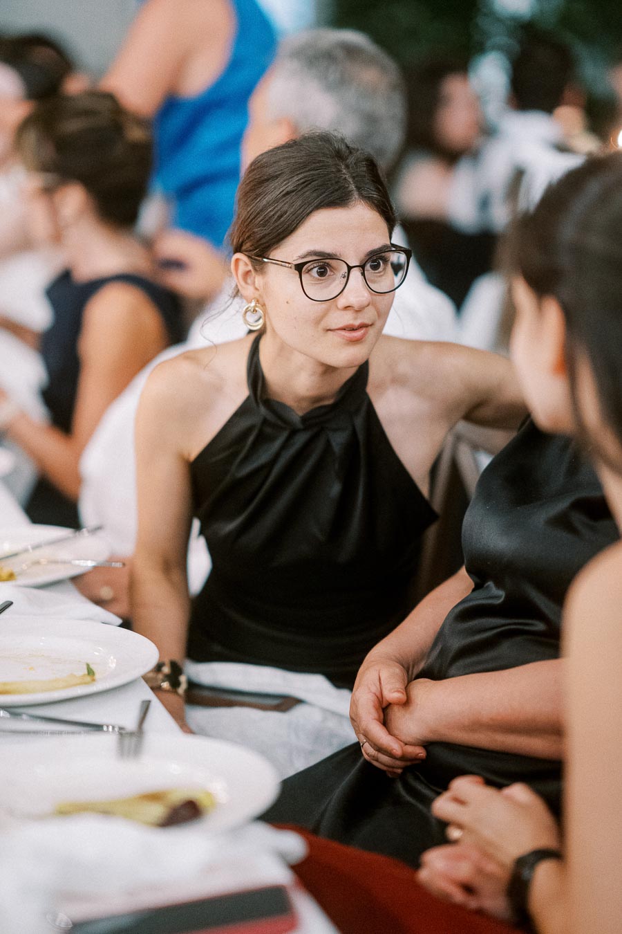 A woman in a black dress and glasses engaged in conversation at a formal dinner event, with people and plates visible in the background.