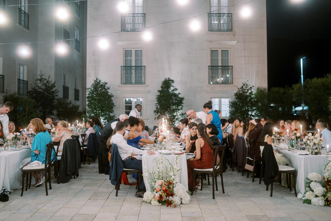 Outdoor evening banquet with elegantly dressed guests seated at long tables, surrounded by decorative flowers and warm lighting, in a courtyard setting.