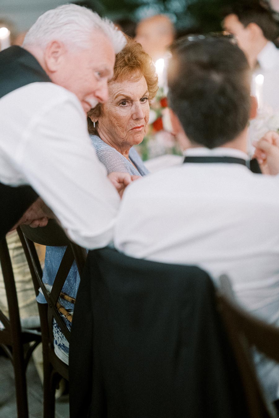 Elderly woman at a formal event engaging in conversation with two men, one standing and one seated, with a softly lit background.