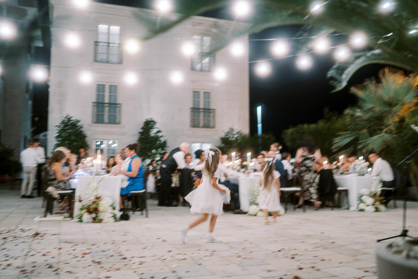 A charming outdoor wedding reception at night, featuring elegantly decorated tables with candles and floral arrangements. Illuminated string lights hang above, creating an enchanting atmosphere as people gather and two young flower girls in white dresses play joyfully in an open courtyard.