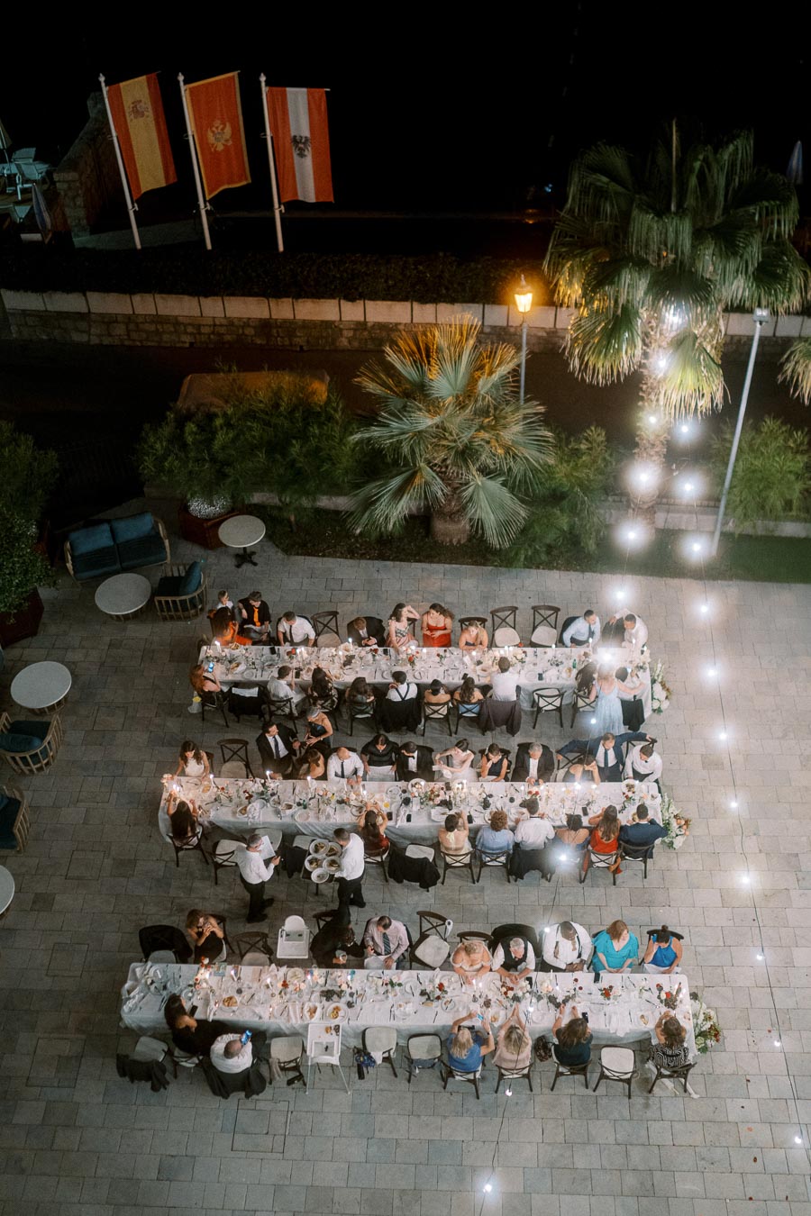 Aerial view of an elegant outdoor dinner party with long tables and guests seated in a lush garden setting, illuminated by string lights, with three national flags displayed in the background.