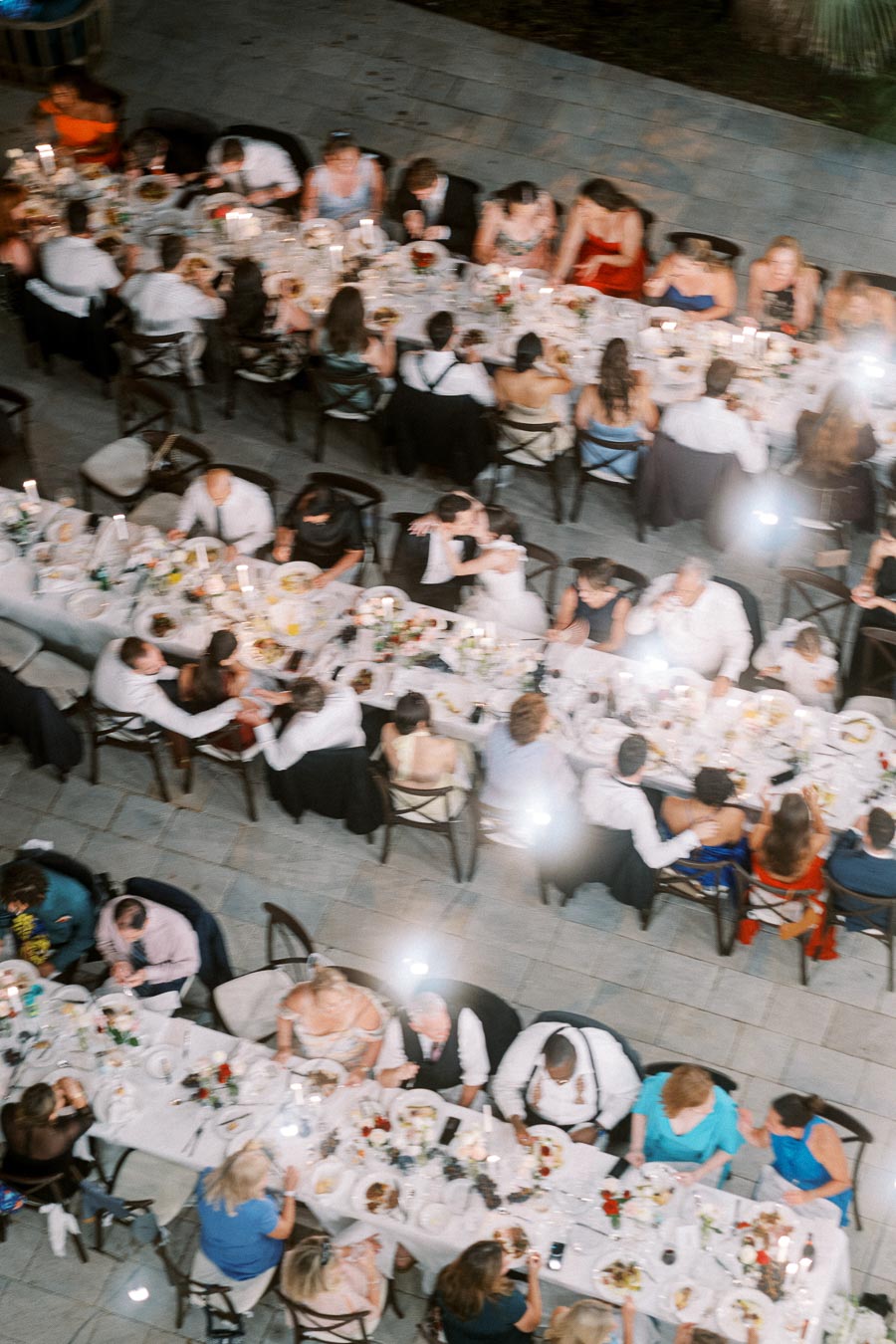 Aerial view of a wedding reception with guests seated at long, elegantly decorated tables, sharing a meal and celebrating under soft lighting.