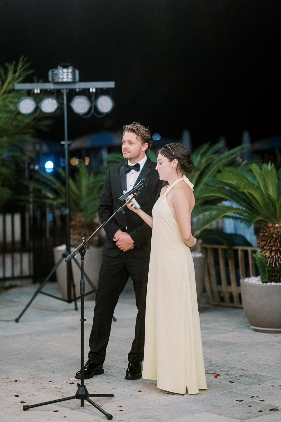 Elegant couple dressed formally, standing outdoors at night, delivering a speech at an event. The woman in a white dress holds a microphone and notes, while the man in a black tuxedo stands beside her. The setting is enhanced by decorative plants and professional lighting in the background.