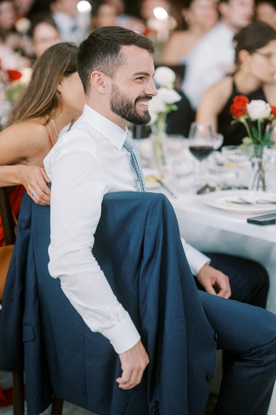 A smiling man in a white dress shirt and blue suit sitting at a dining table during an elegant event, with tables set with wine glasses and flowers in the background.