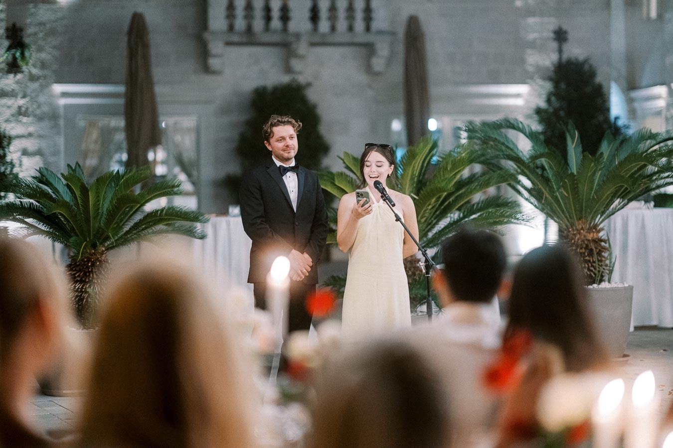 A woman in a white dress gives a speech at a wedding reception, standing beside a man in a tuxedo. They are in a beautifully decorated outdoor venue with lush green plants and elegant table settings.