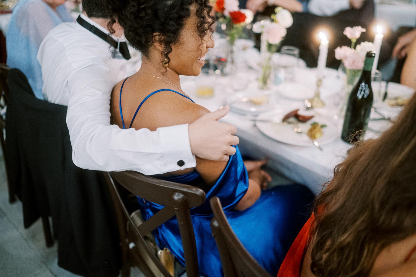 Couple sitting together at a formal dinner table, elegantly dressed with a man in a white shirt embracing a woman in a blue dress, surrounded by decorated table settings and flowers.