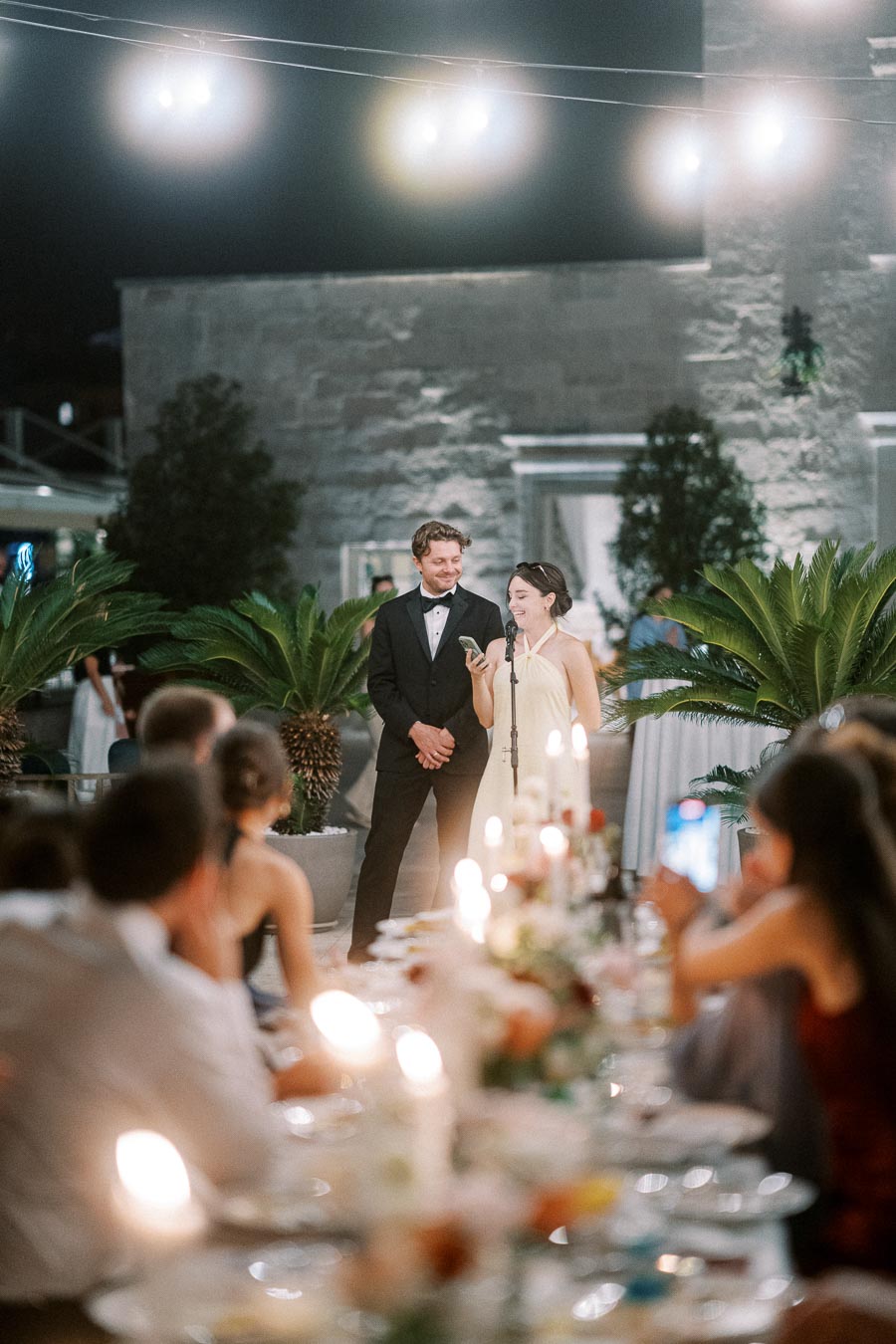 A well-dressed couple gives a speech at an outdoor wedding reception, surrounded by guests seated at elegantly decorated tables with candles and flowers at night.