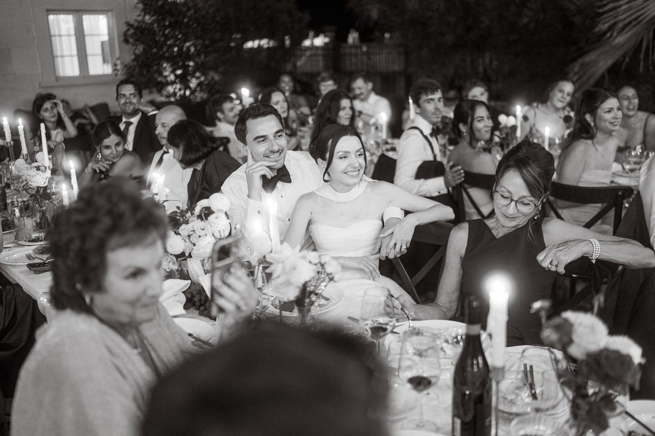Black and white photograph of a joyful wedding reception with guests seated around candlelit tables, featuring a smiling bride and groom at the center, surrounded by friends and family enjoying the celebration.