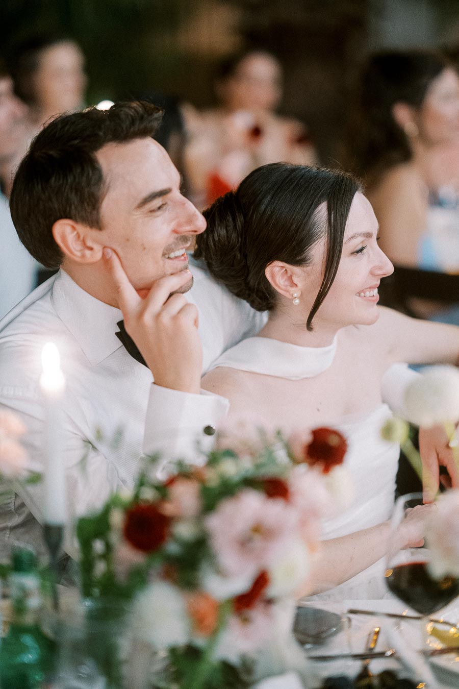 A smiling bride and groom sharing a joyful moment at their wedding reception, surrounded by blurred guests and elegant floral decorations.