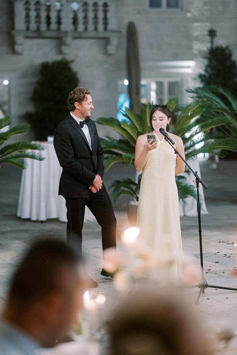 Man in a tuxedo and woman in an elegant dress giving a toast at a formal event with a microphone, surrounded by elegant decor and ambient lighting.