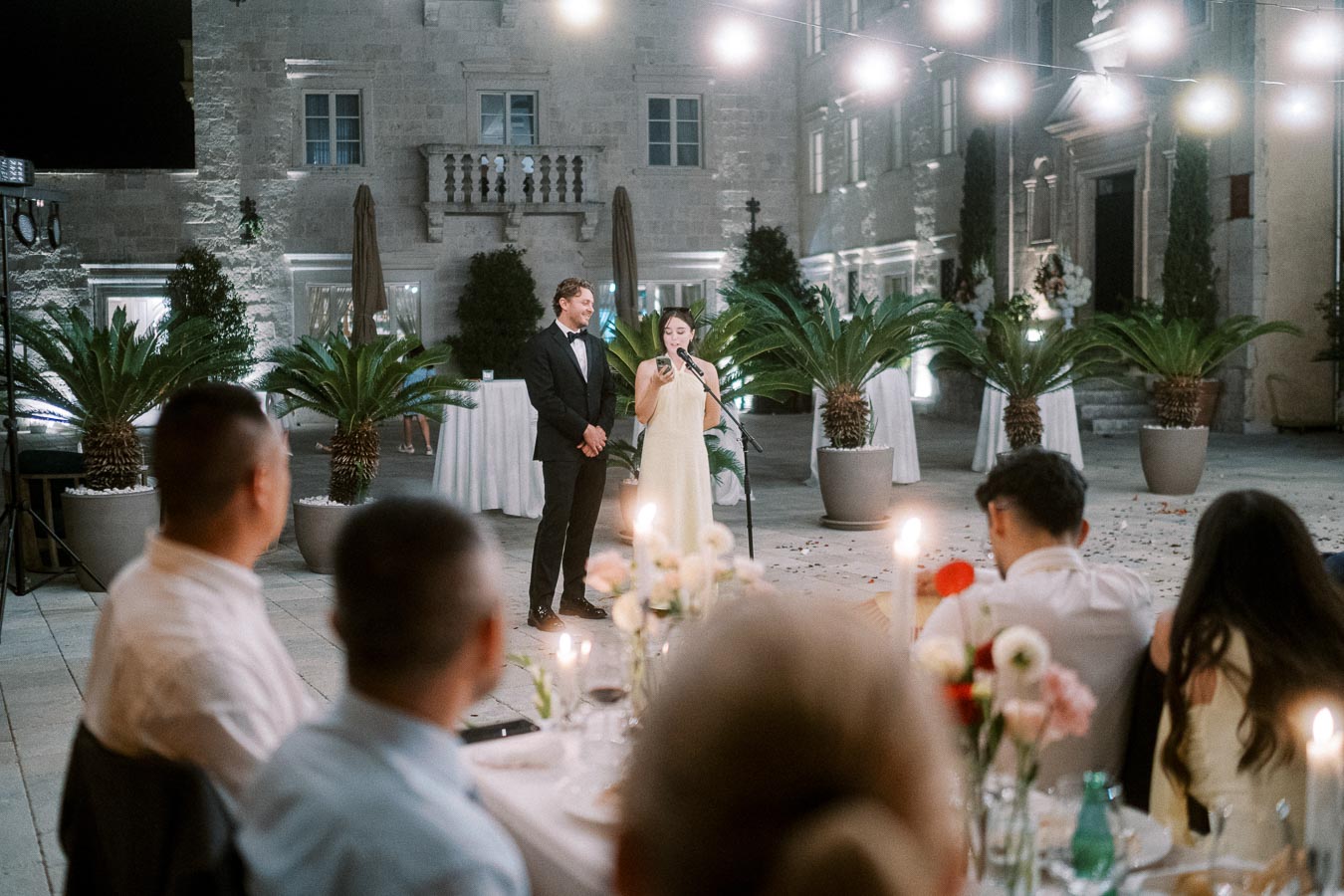 A couple giving a speech at an elegant wedding reception in an outdoor courtyard, with guests seated at candle-lit tables, surrounded by lush potted plants and decorative lights hanging overhead.
