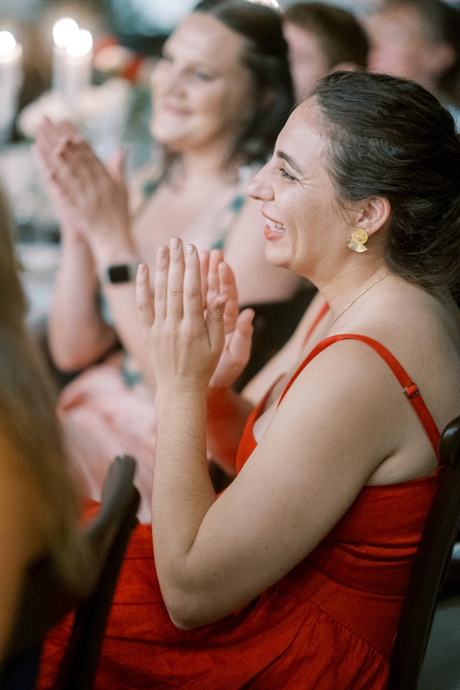 A woman in a red dress clapping and smiling at an indoor event, with blurred background and candlelight ambiance.