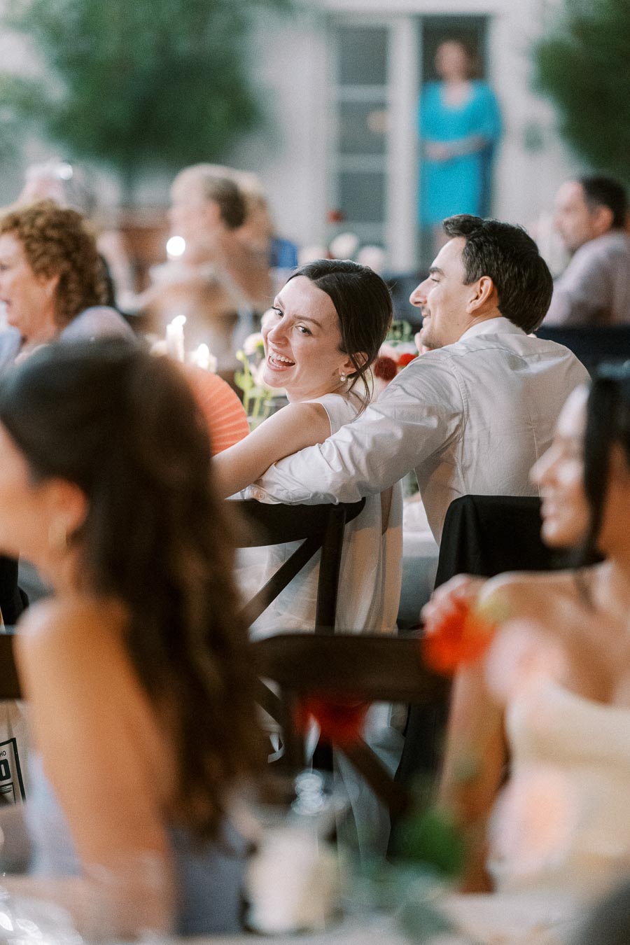 Smiling couple enjoying an outdoor wedding reception with guests seated around tables, decorated with candles and colorful flowers.