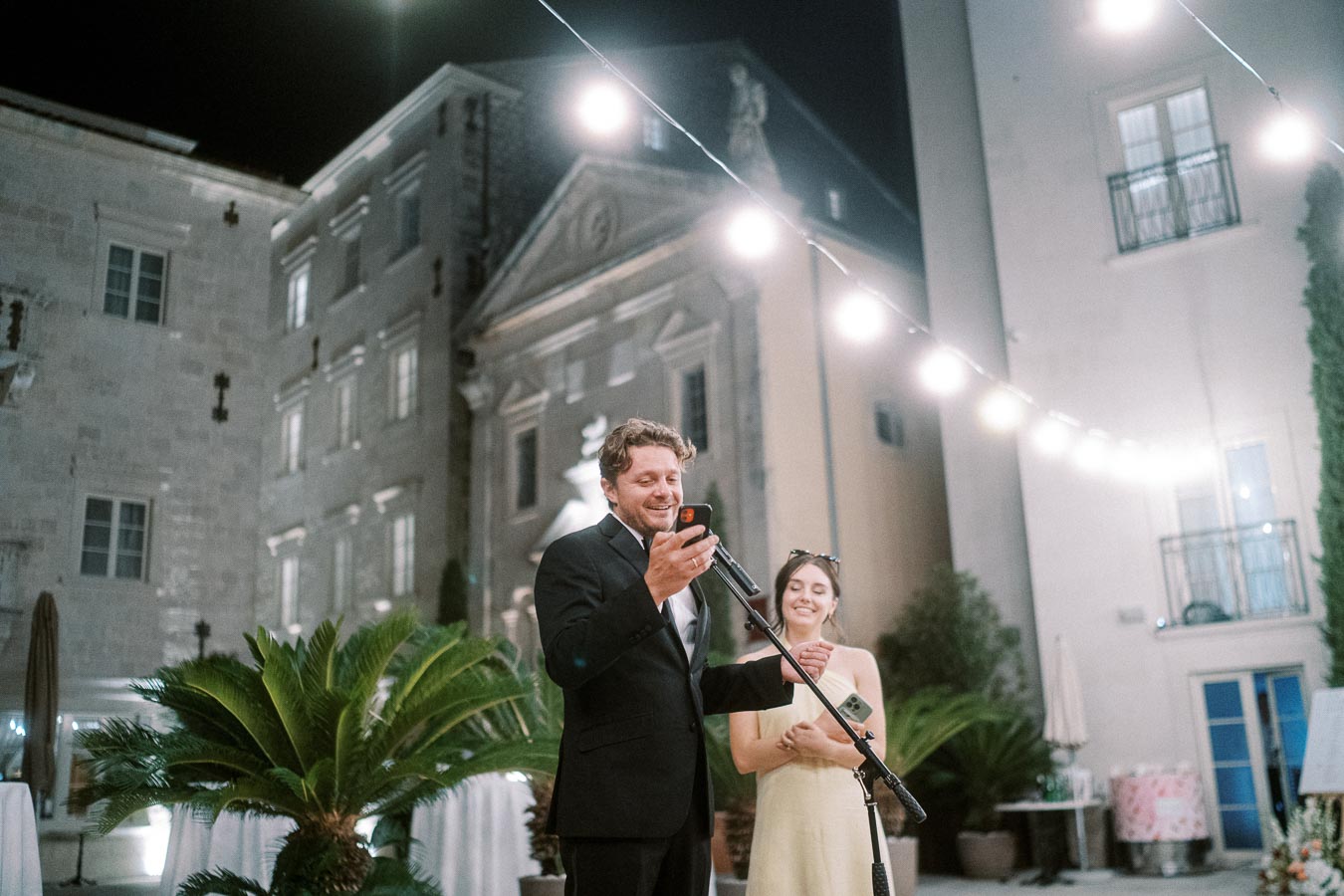 Wedding guests giving a speech outdoors at night, with festive string lights and historic architecture in the background.