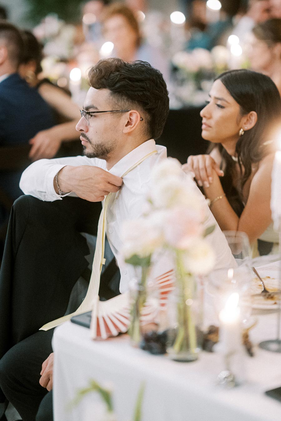 A man wearing glasses and adjusting his tie while a woman sits beside him at a formal event, with blurred guests and a candlelit setting in the background.