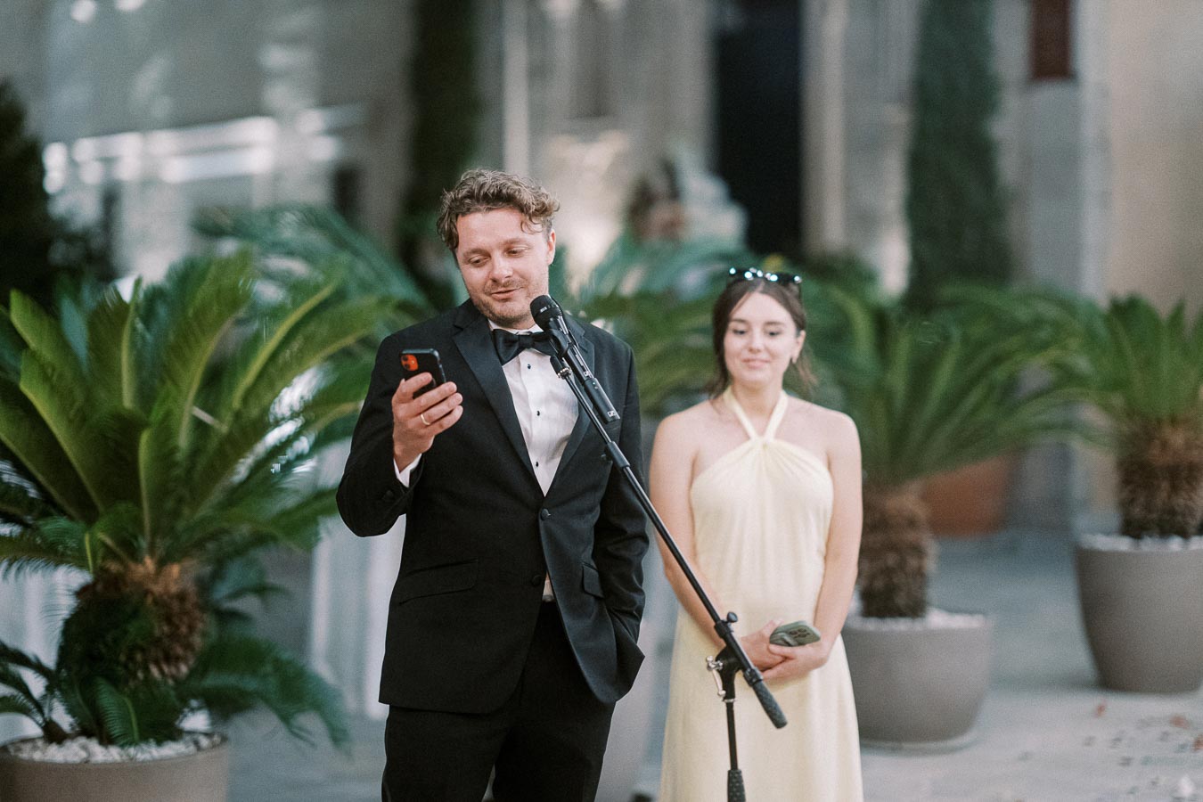 Man in a tuxedo giving a speech at an outdoor event, standing next to a woman in a yellow dress with potted plants in the background.