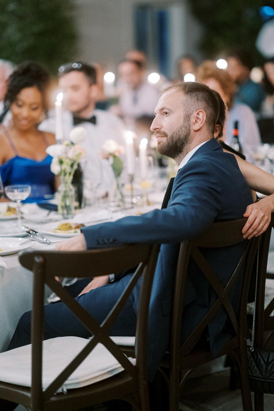 A well-dressed man in a blue suit sits attentively at an elegantly set dinner table during a formal event, surrounded by guests with candles and flowers as centerpiece decor.