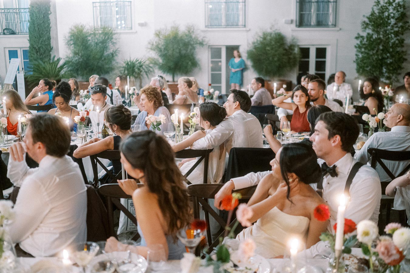 Elegant wedding reception with guests seated at candlelit tables, adorned with floral centerpieces in an outdoor courtyard setting.