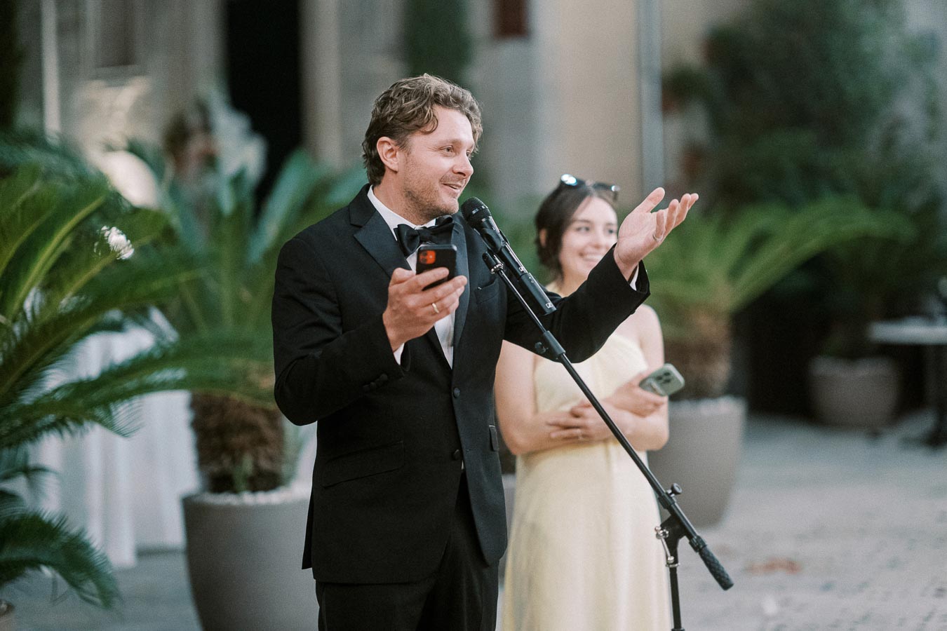 Man in a black tuxedo delivering a speech at an outdoor event, holding a smartphone and gesturing, with a woman in a yellow dress smiling in the background.