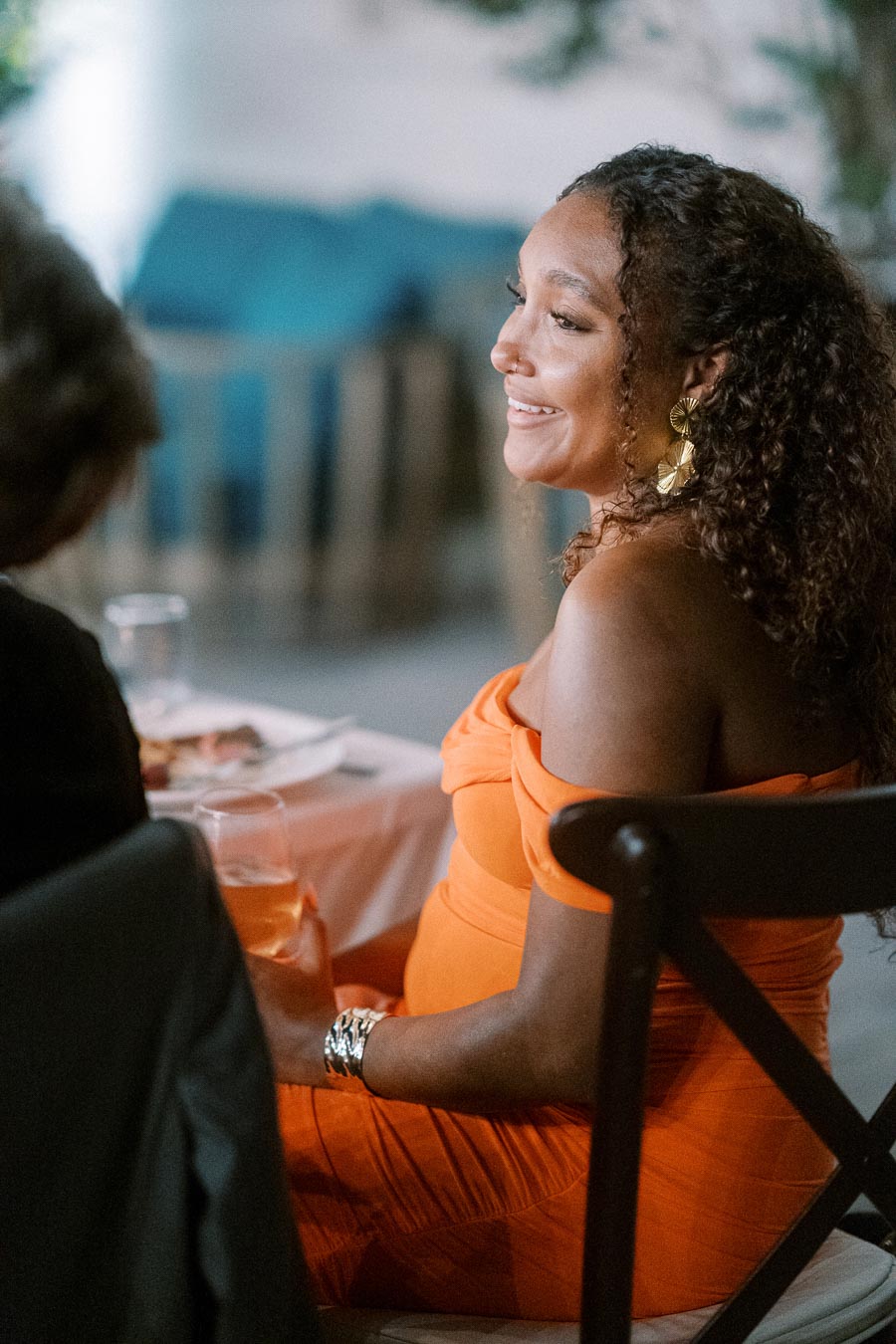 A woman in an orange dress with curly hair and gold earrings smiling during a social gathering at a restaurant.