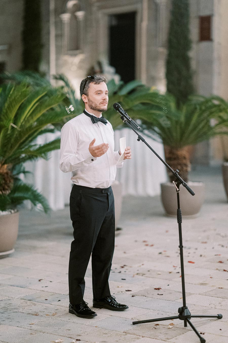 A man in formal attire gives a speech outdoors, standing near a microphone with lush green plants in the background.