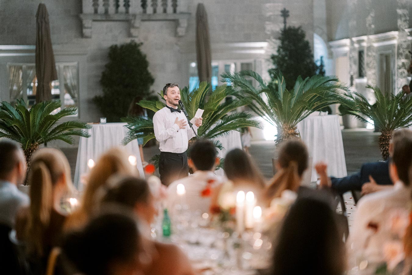 Man giving a speech at an elegant outdoor event with candlelit tables and decorative greenery, creating a warm and inviting atmosphere.