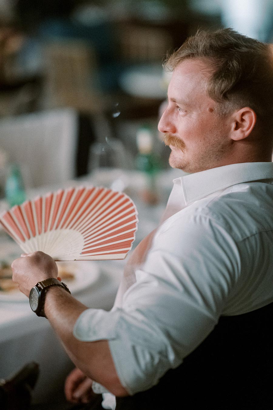 Man seated at a dining table holding a decorative handheld fan, dressed in a white shirt with a watch on his wrist, setting an elegant and relaxed atmosphere.