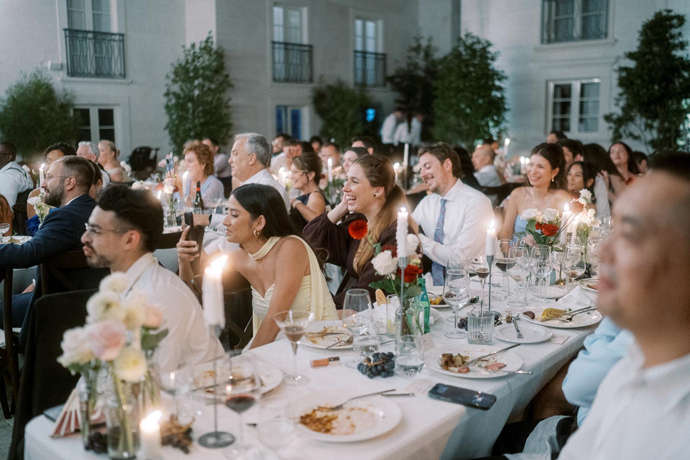 A lively wedding reception with guests seated at elegantly decorated tables, enjoying the celebration with lit candles and floral centerpieces. The guests appear engaged and joyful in a warmly lit venue.