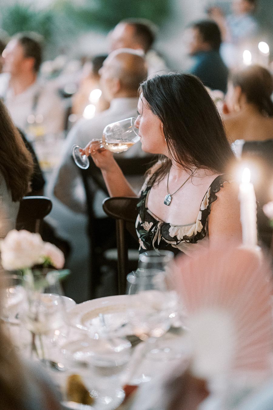 A woman in a floral dress enjoys a glass of wine at an elegant candlelit dinner party, surrounded by blurred guests and soft lighting.