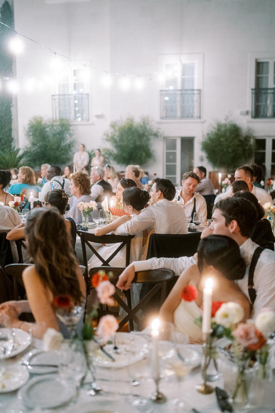 Outdoor wedding reception with elegantly dressed guests seated at decorated tables, under string lights, enjoying a festive evening ambiance.