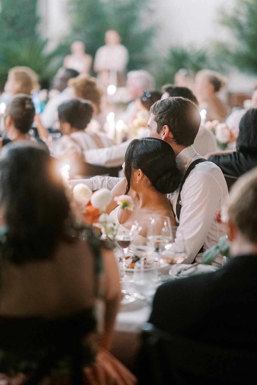 A couple sitting closely at an elegantly decorated wedding reception, surrounded by guests and soft candlelight.