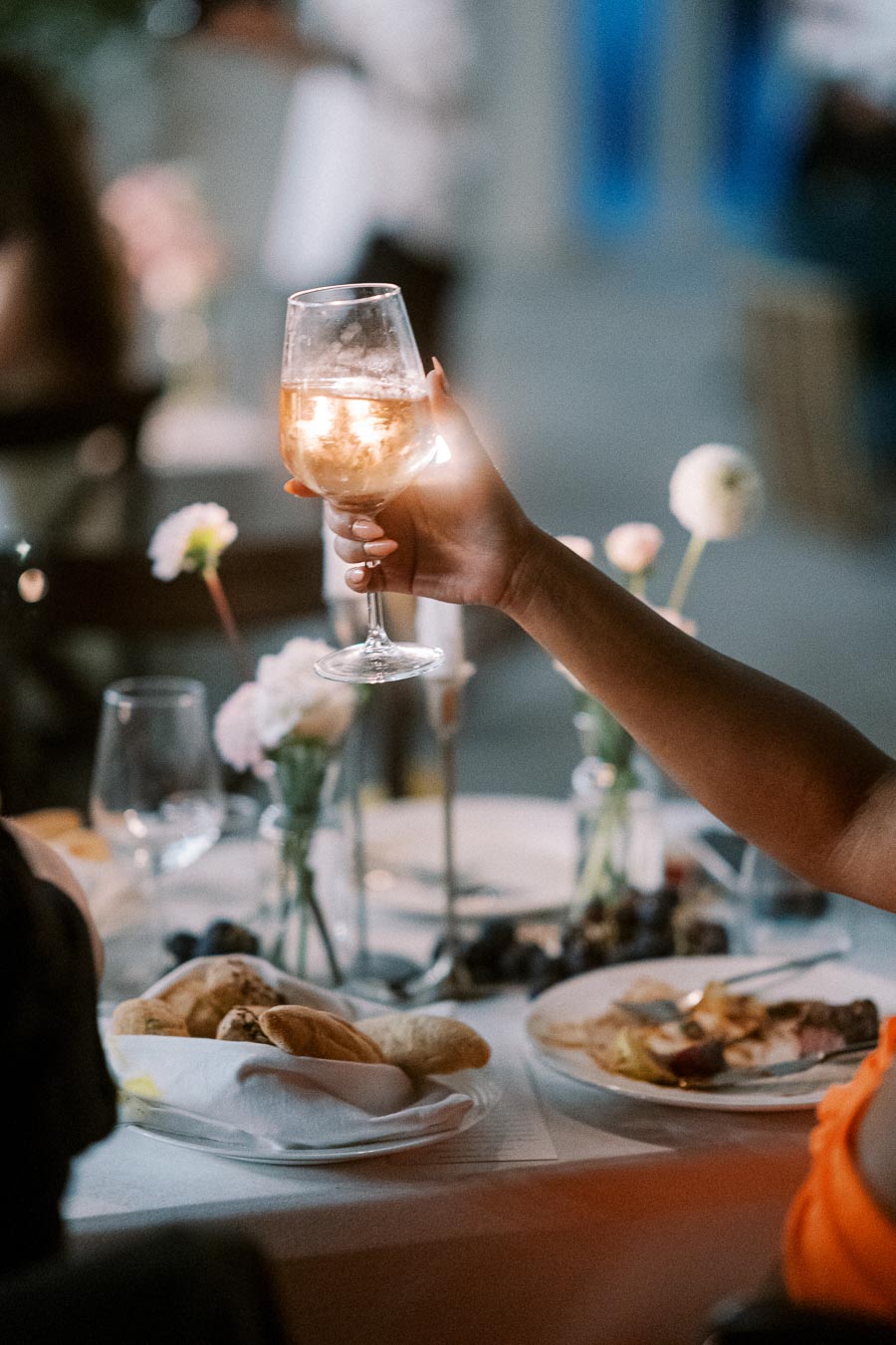 A hand elegantly holding a glass of white wine at a dimly lit dinner party, with a table set with plates of food, fresh bread, grapes, and floral arrangements, creating a warm and inviting ambiance.