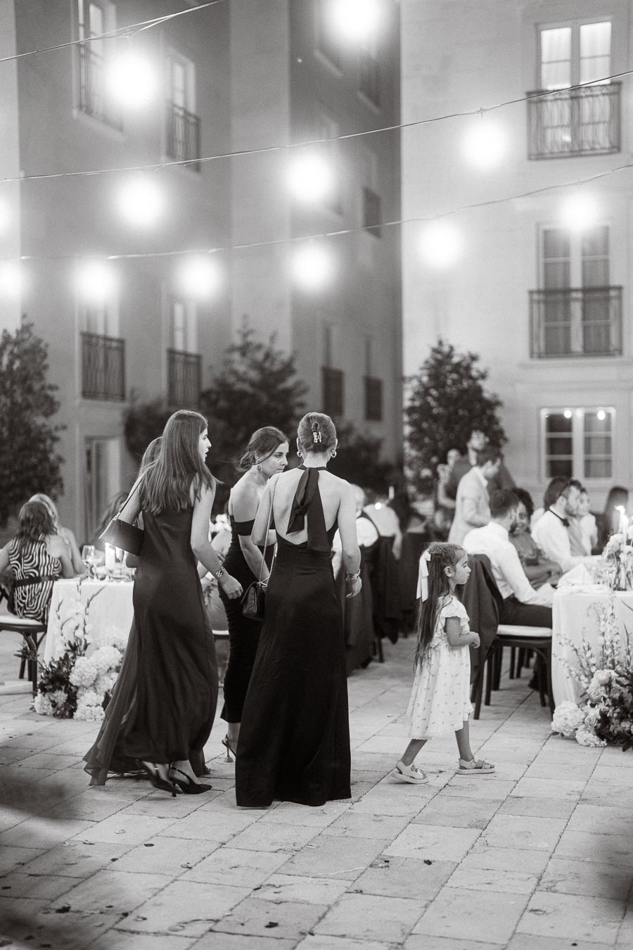 A black and white photo of an elegant outdoor evening event, featuring people in formal attire, including women in dresses and a child in a white dress, with string lights illuminating the venue.