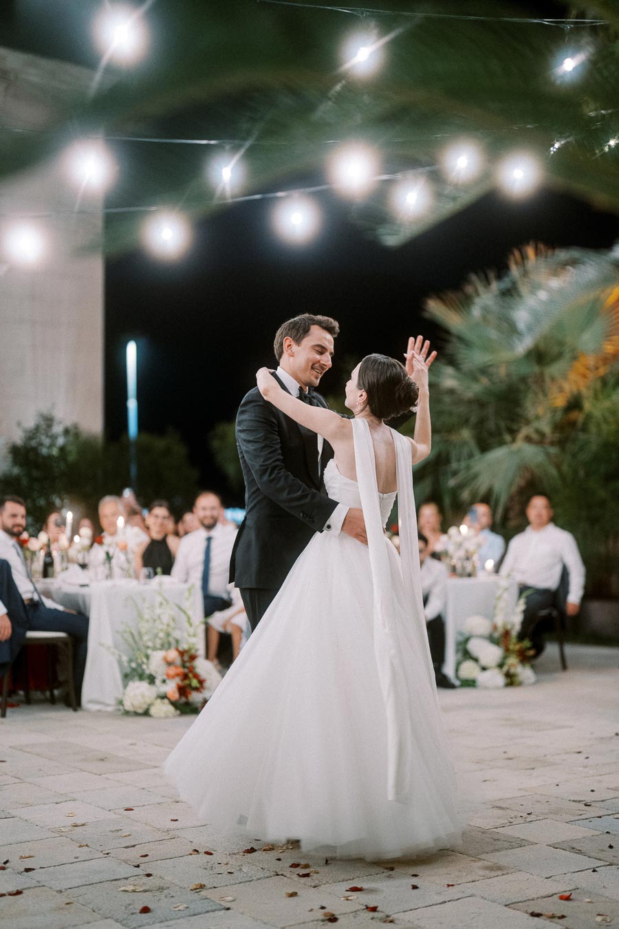 Newlywed couple sharing a romantic first dance under string lights at an outdoor evening wedding reception, surrounded by guests seated at decorated tables.