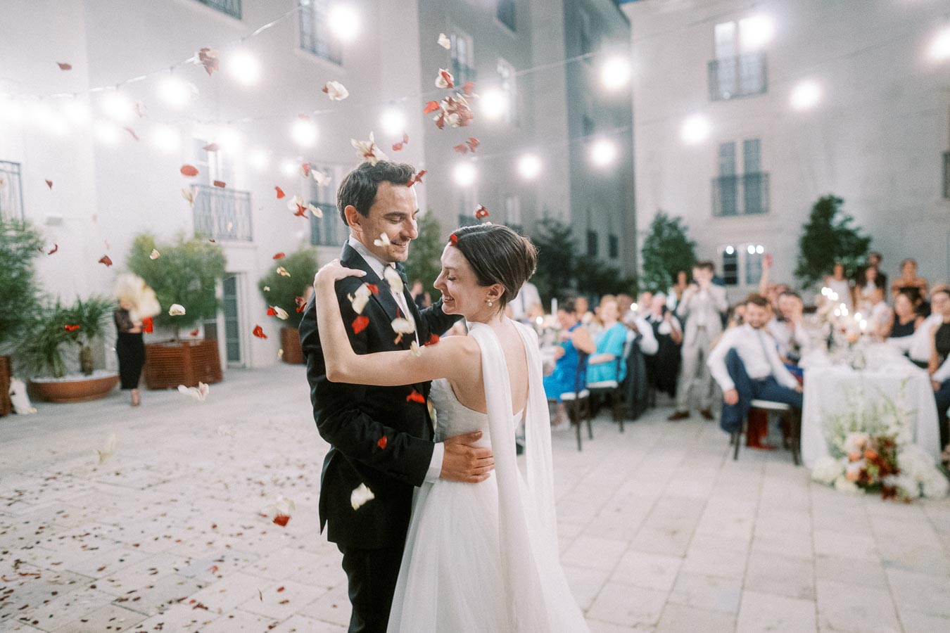 Romantic outdoor wedding reception with bride and groom sharing a first dance under string lights, surrounded by guests and showered with rose petals.