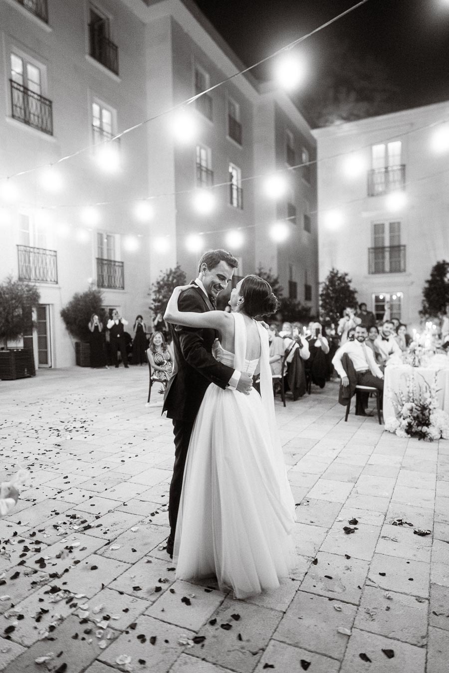 A bride and groom share a romantic first dance under string lights in a courtyard setting, surrounded by guests in elegant attire.